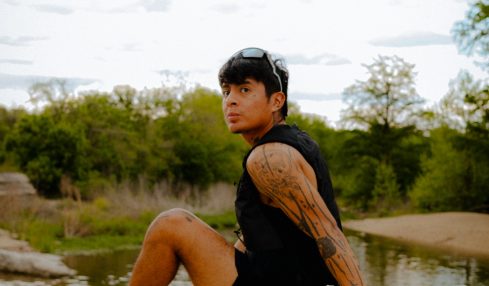 Person sitting on a rock near water with trees and cloudy sky in background, wearing black clothing and colorful shoes.