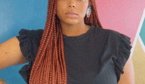 Young woman with long braided hair wearing a black top with ruffled sleeves, standing against a colorful geometric background.