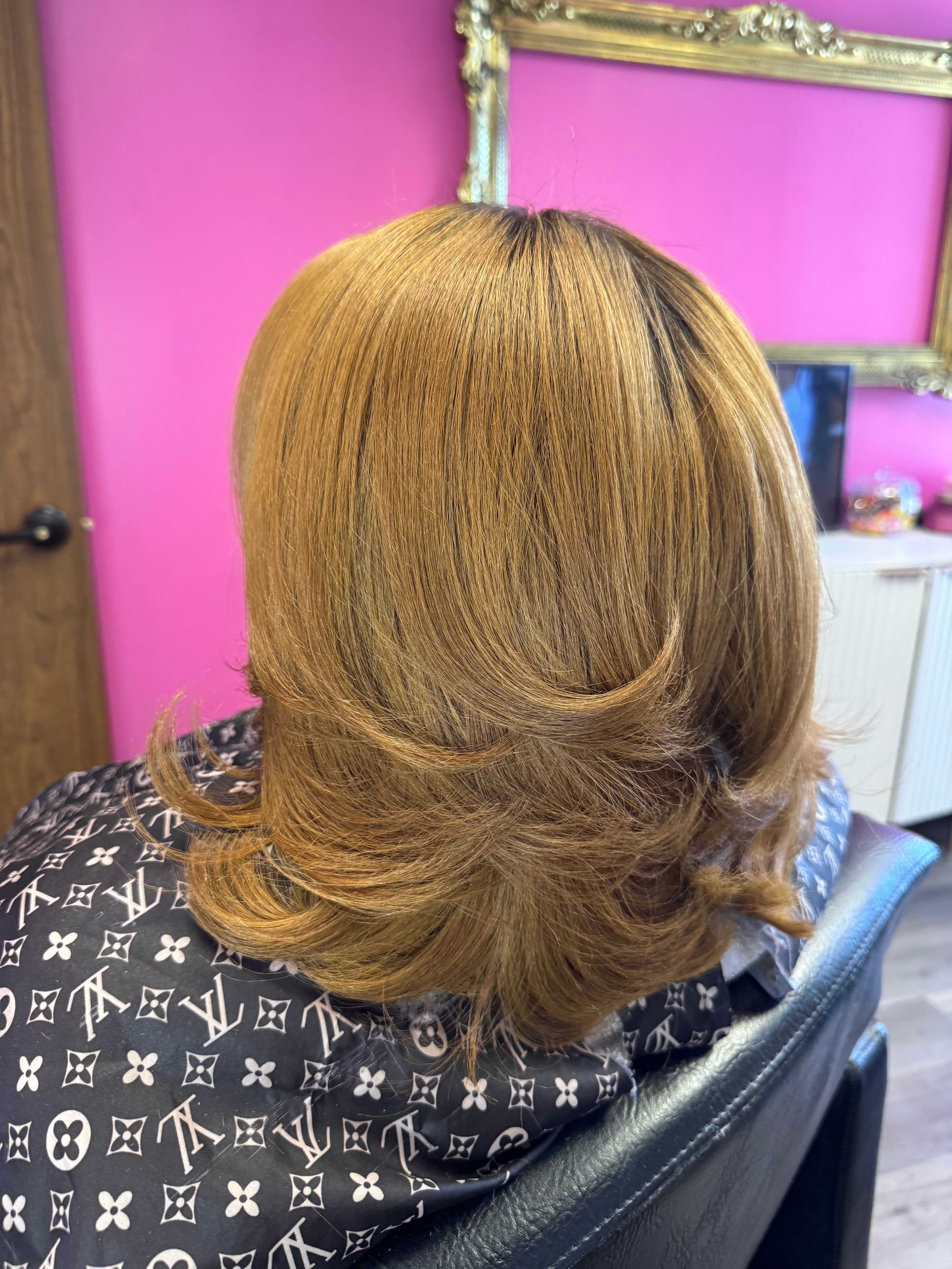 Side view of a person with shoulder-length, light brown hair, sitting in a salon chair with a pink wall background.