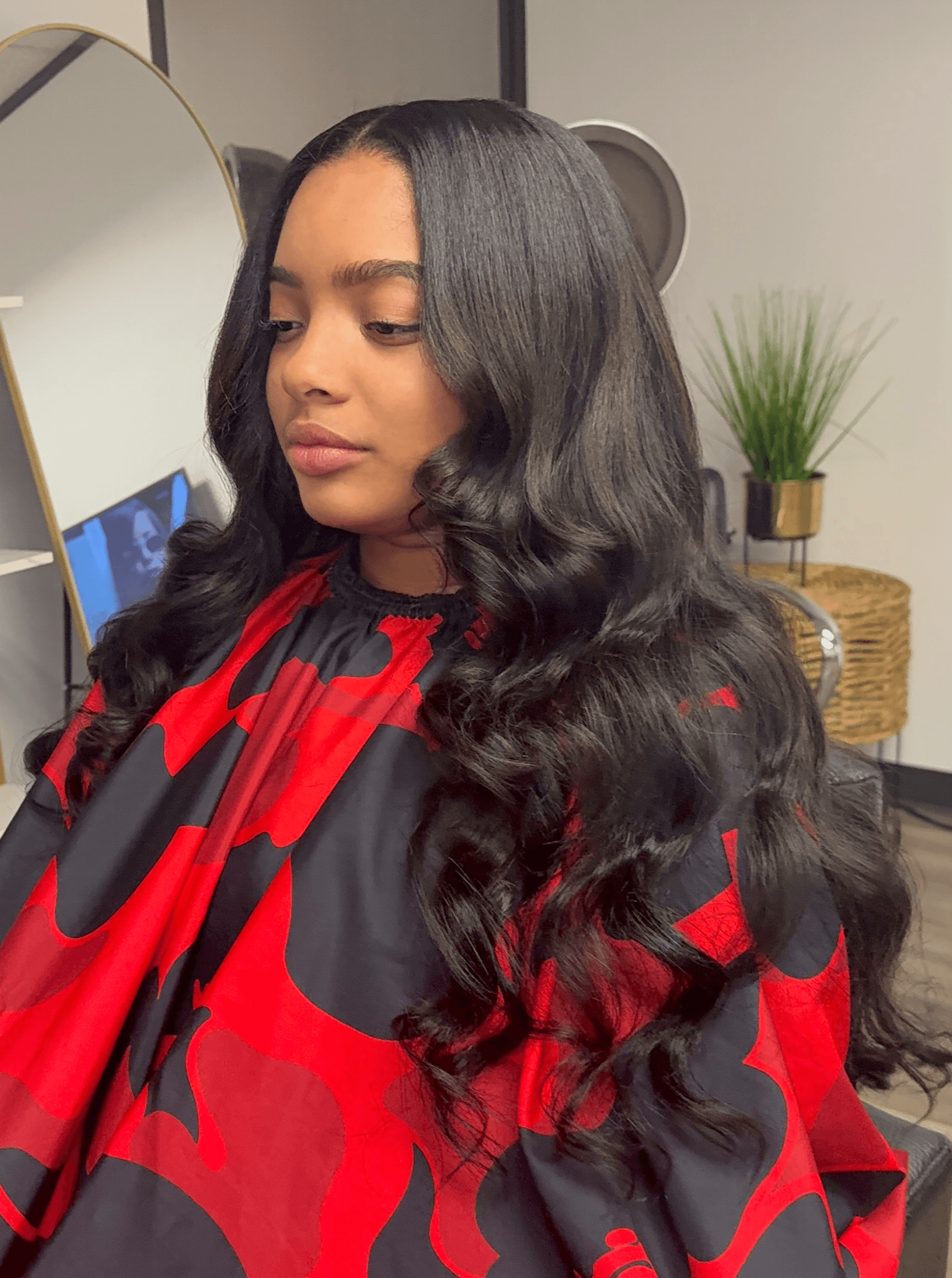 Young woman with long, wavy black hair wearing a red and black patterned top, looking to the side indoors.