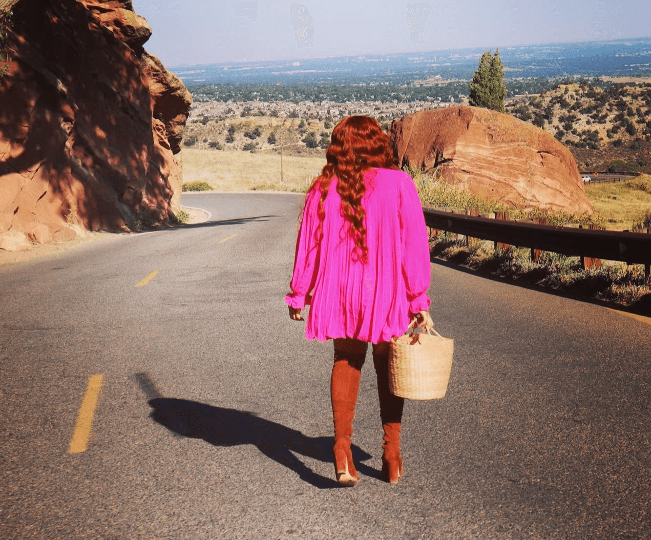 Person with long red hair walking on a winding road holding a basket, landscape with rocks and trees in background