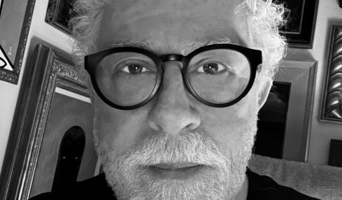 Portrait of an older man with curly white hair, beard, and glasses, in a room with framed pictures on the wall.