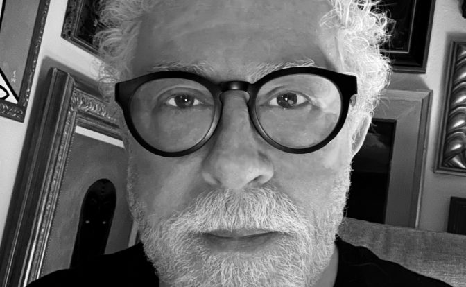 Portrait of an older man with curly white hair, beard, and glasses, in a room with framed pictures on the wall.