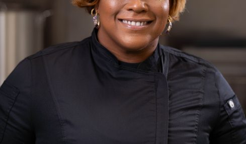 Smiling woman with short, wavy blonde hair wearing a black top, standing indoors.
