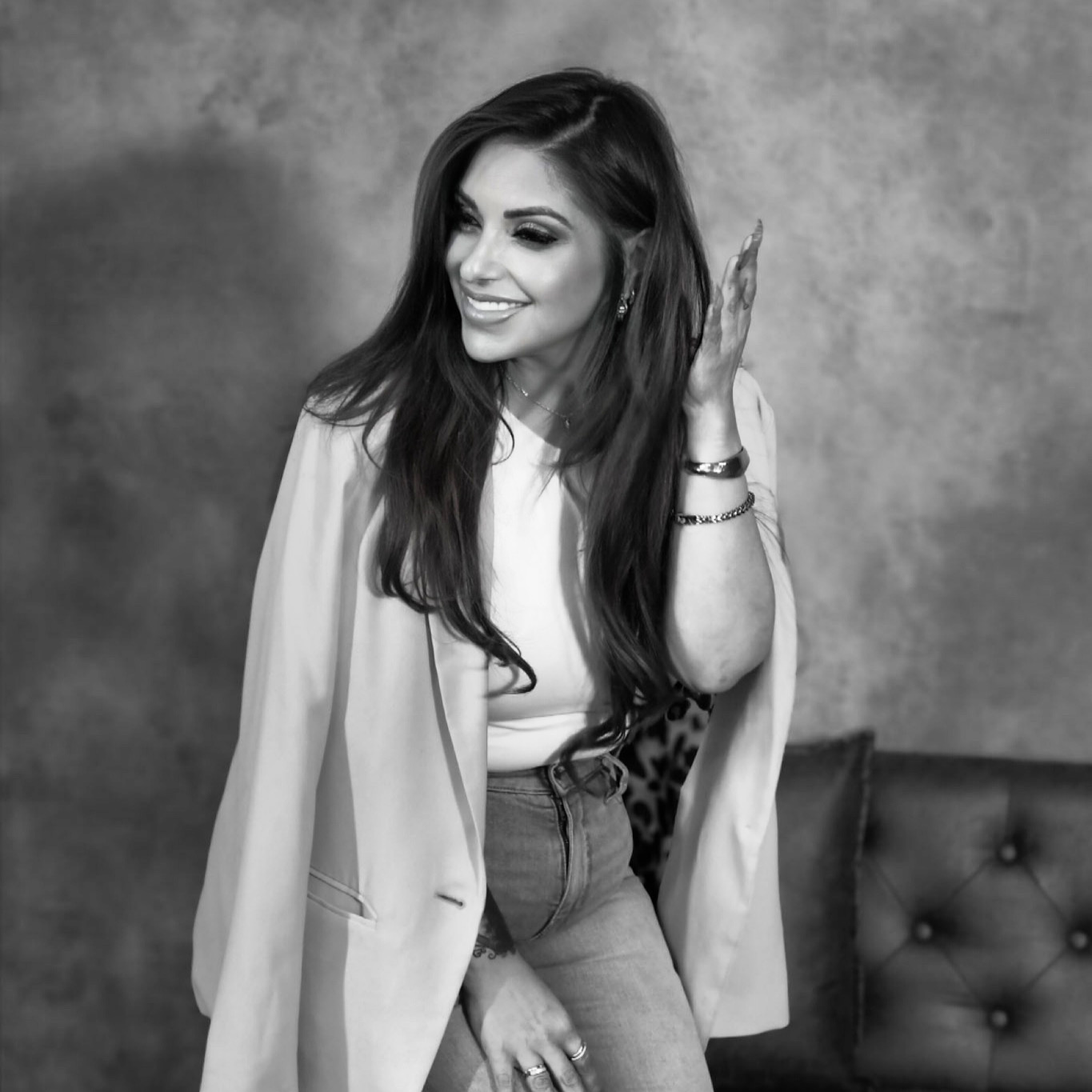 Smiling woman with long hair, wearing a blazer and jeans, sitting on a sofa, touching her hair, in black and white.