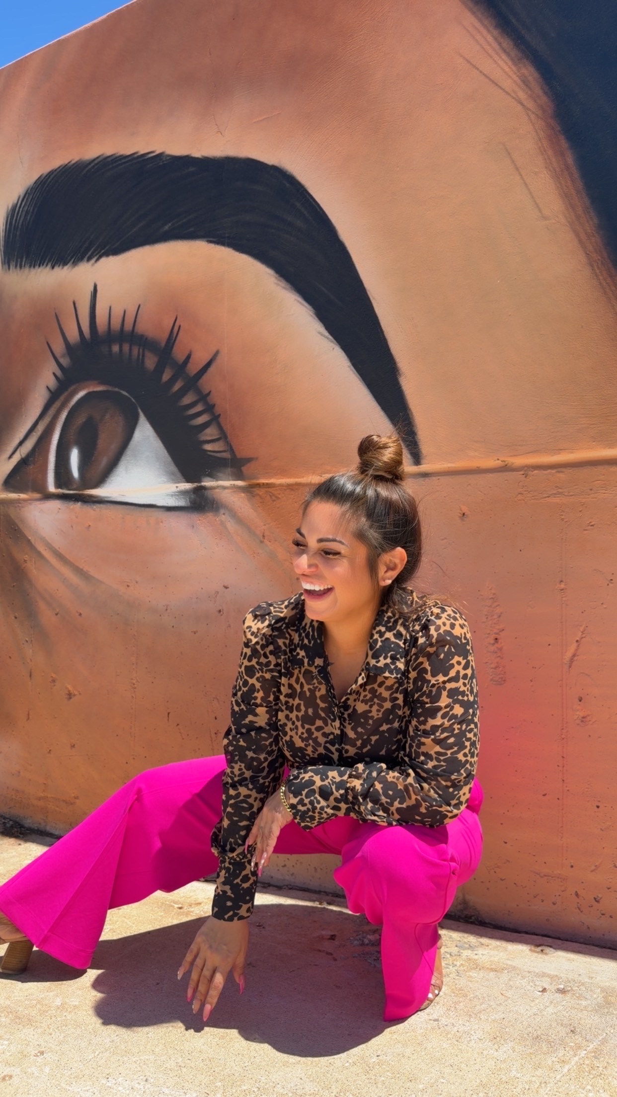 Young woman with a bun, smiling, wearing a leopard print top and bright pink pants, sitting on the ground in front of a large mural of a woman's eye.