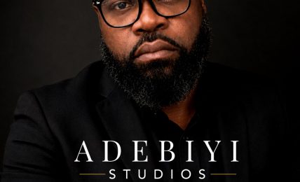 Portrait of a man with glasses and a beard, wearing a black shirt, against a dark background.